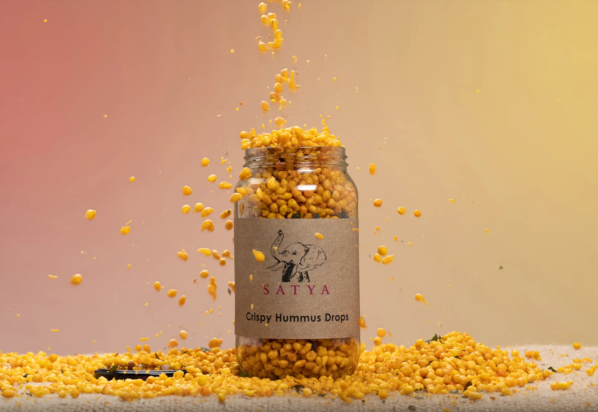 Golden hummus drops pouring into a Satya jar against a warm pink-to-gold ground.