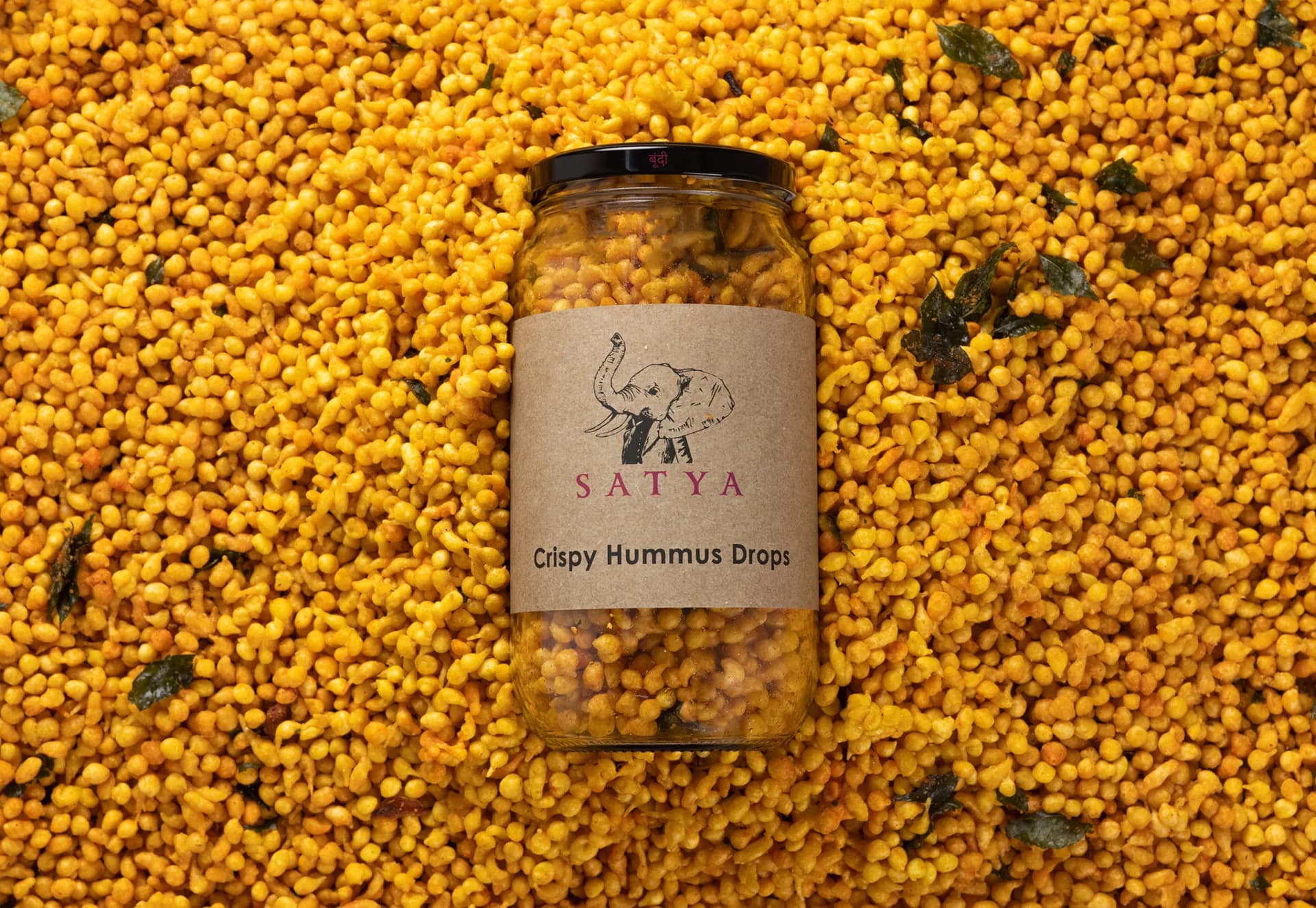 A Satya jar of Crispy Hummus Drops lying in a bed of hummus drops.
