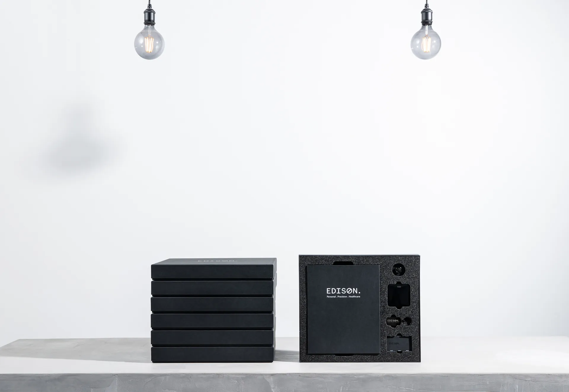 A stack of black Edison boxes beside an open kit, under two hanging bulbs.