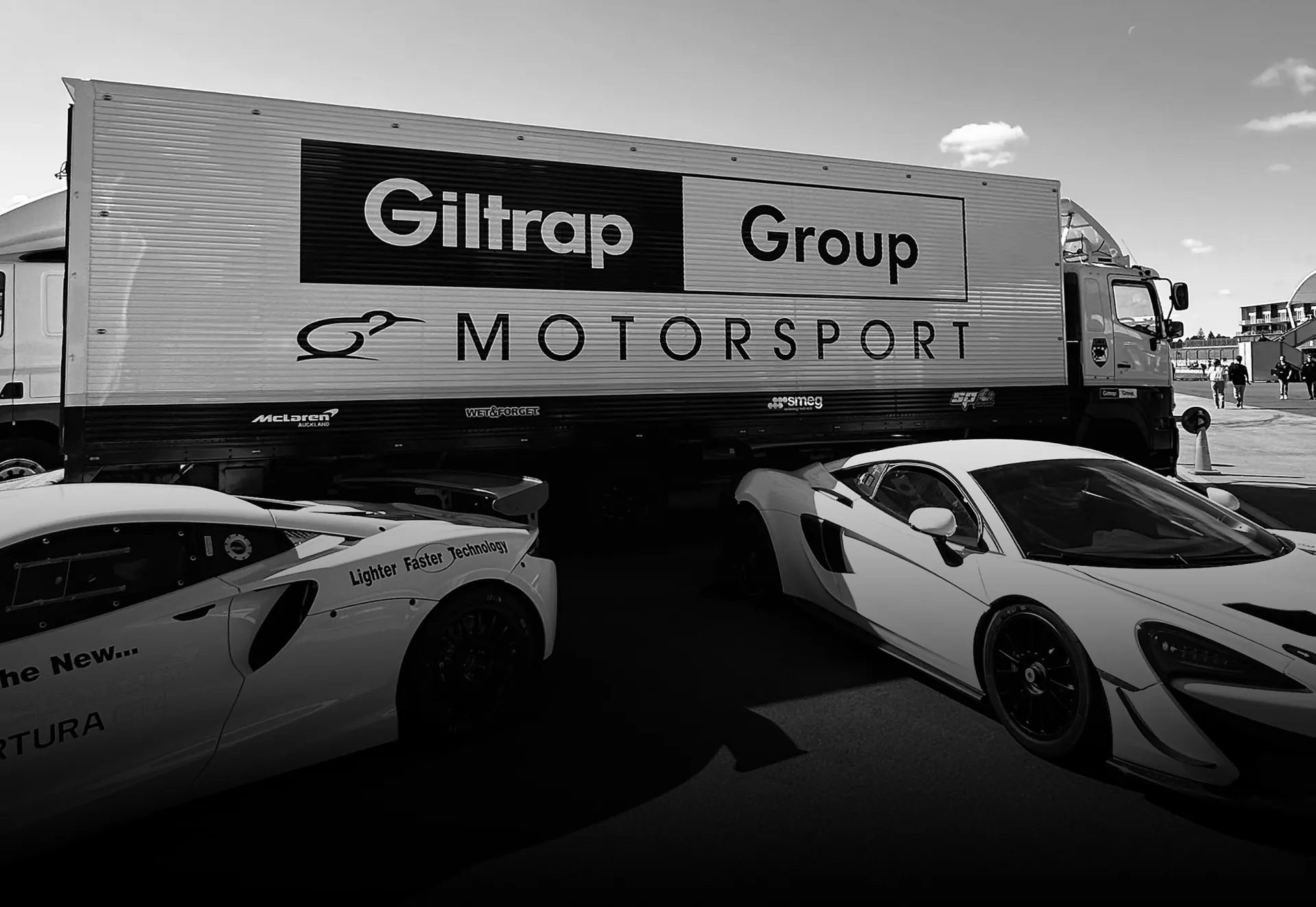 A Giltrap Group Motorsport transporter behind two white McLarens, in black and white.