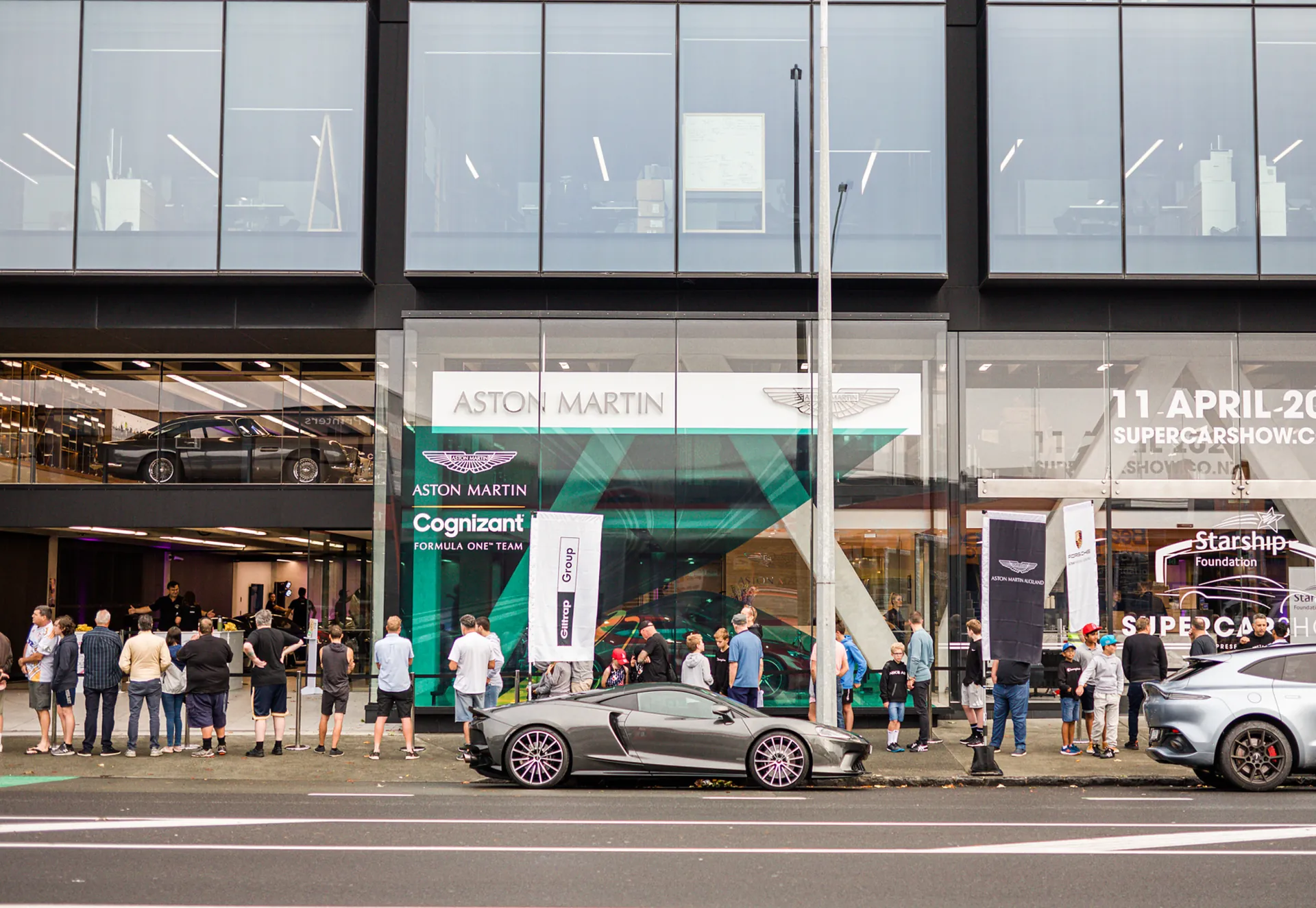Visitors queuing outside the Supercar Show at the Aston Martin showroom, a supercar at the kerb.