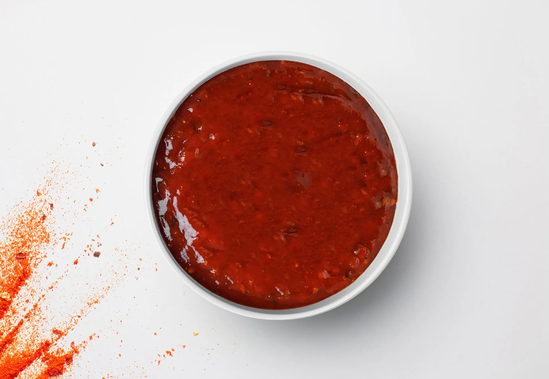 A white bowl of hot sauce from above, red chilli powder beside it.